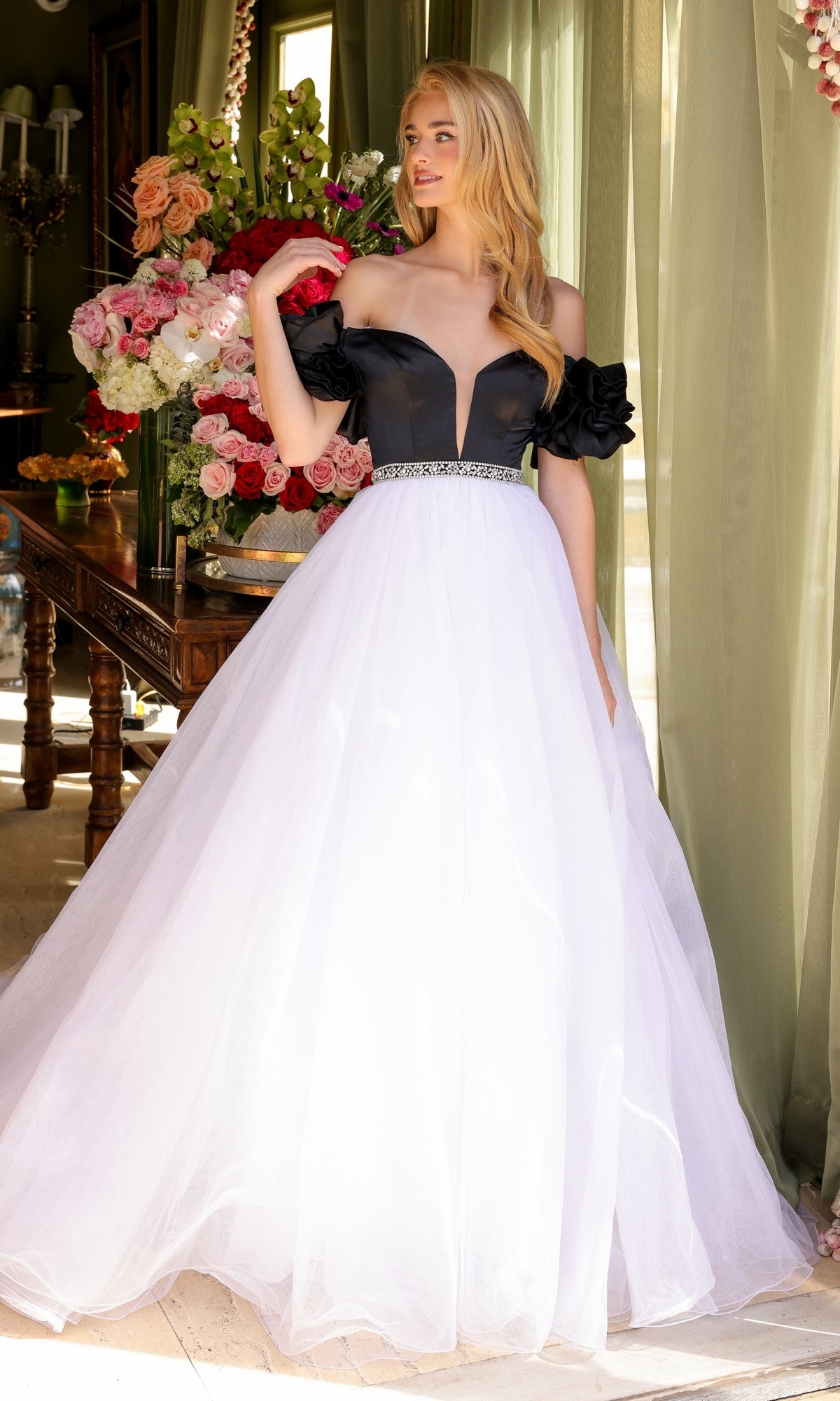 A woman stands indoors near green curtains and a floral-topped table, wearing the Ava Presley Long Prom Dress 39581—an off-the-shoulder black and white A-line ball gown with a full tulle skirt.